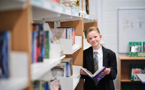 White boy in smart school uniform leaning against a large bookshelf reading a hardback book