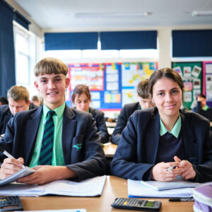 WA14 Boy and girl, aged approximately 16, sat behind a school desk cmiling whilst doing mathematics-based work with calculators.